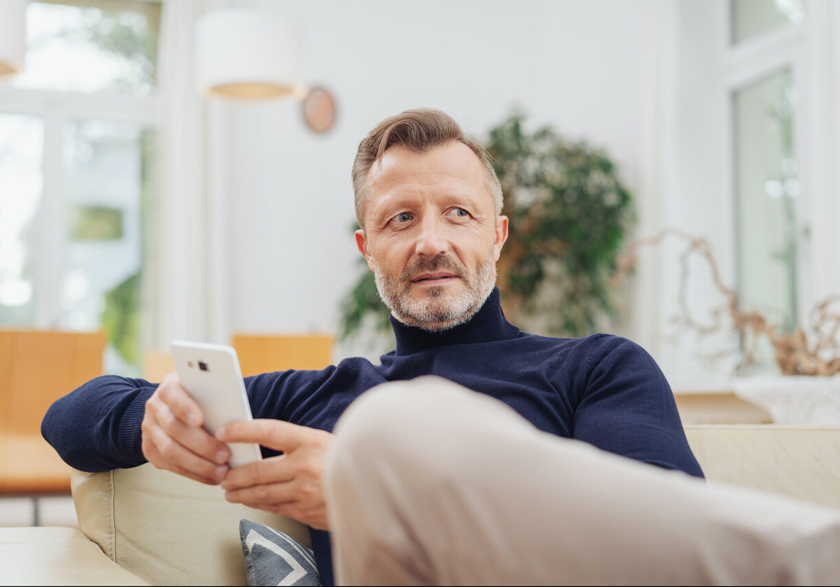 Trendy bearded man with a speculative expression relaxing on a couch at home holding a mobile phone and looking pensively to the side